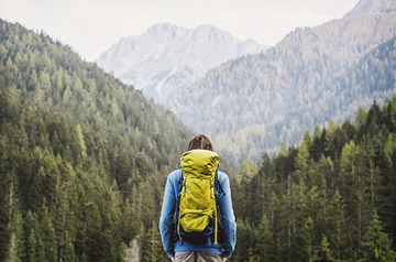 person carrying a yellow backpack looks at forested mountain