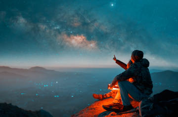 Hiker seated on a mountaintop overlooking water at night.